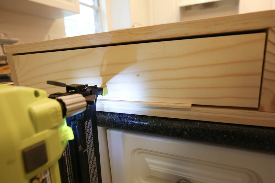 Attaching the drawer faces to the makeup vanity with a brad nailer from the front using a paint stick stirrer to hold in place