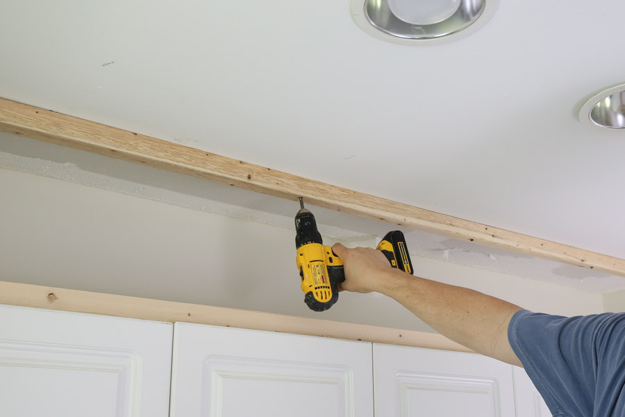 use screws and drill to attach wood board into ceiling studs above cabinets