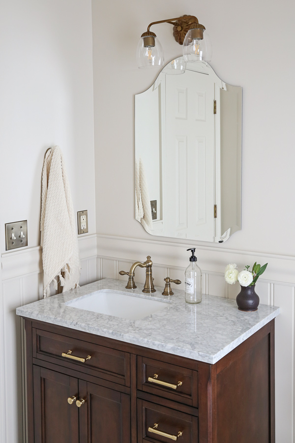 bathroom makeover with beadboard wainscoting, chair rail, marble tile flooring, brass faucet, and frameless arch mirror and wood vanity